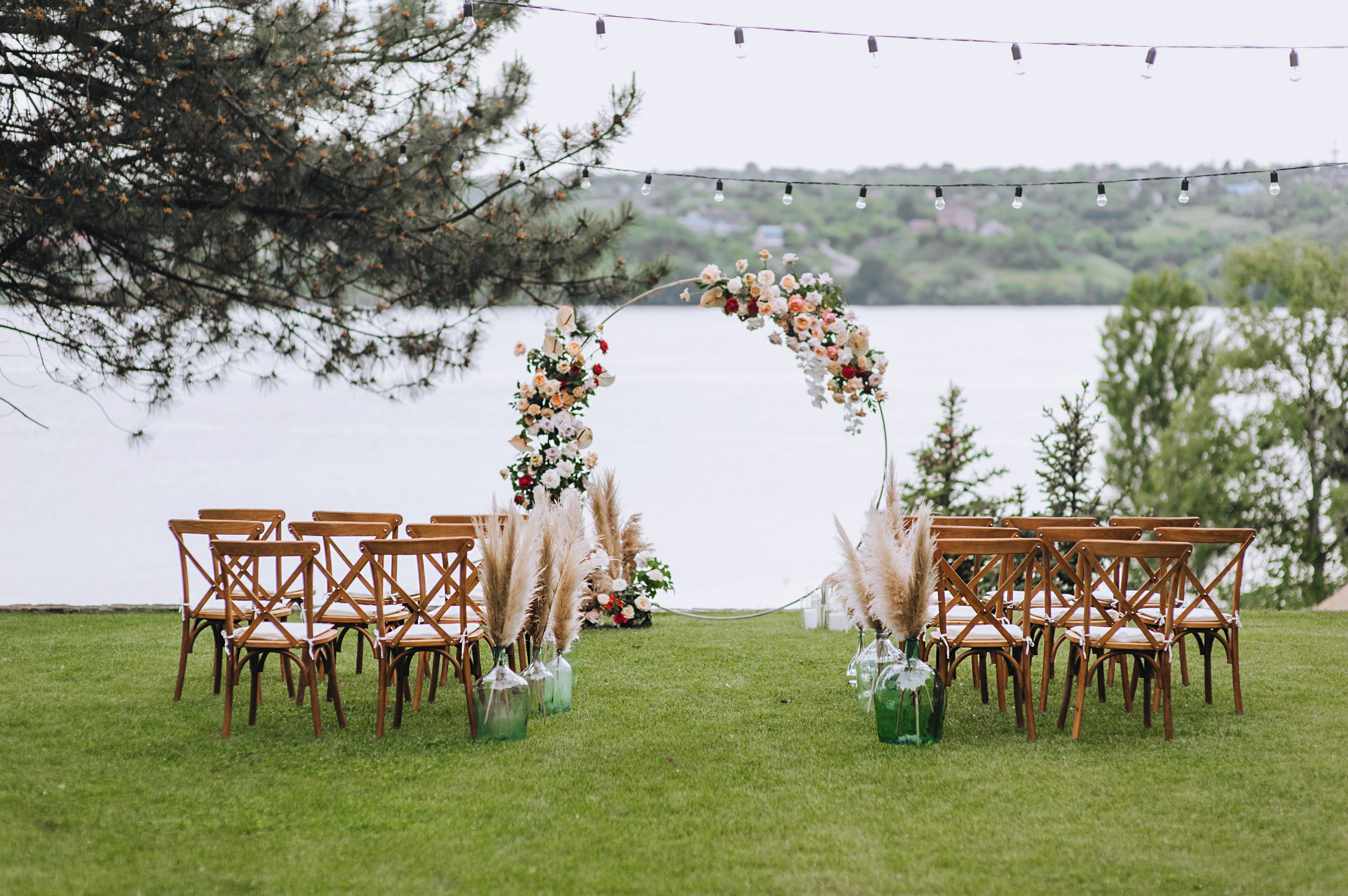 Wedding arch decorated with flowers, wooden chairs stand on green grass against the background of the river on a sunny day. Outdoor ceremony. A place to celebrate. Photography, concept.